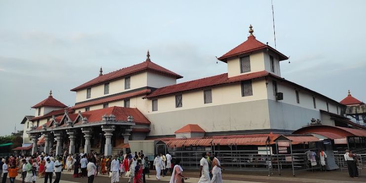 A bustling pilgrimage site in Dharmasthala, Karnataka, with a large white and red building and people walking around.