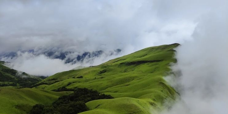 A scenic green hill with clouds and grass, part of the challenging Netravati Peak trek in India.