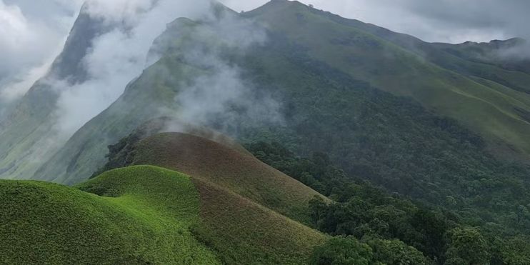 A breathtaking mountain range with lush green hills, fluffy clouds, and a challenging trek to Netravati Peak summit.