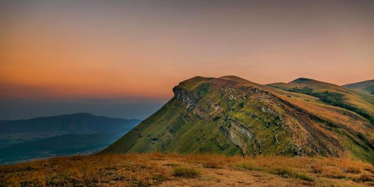 The sun sets over Kemmangundi Peak's mountain range, grass, and hill. Ideal months to visit are September through March for pleasant weather.