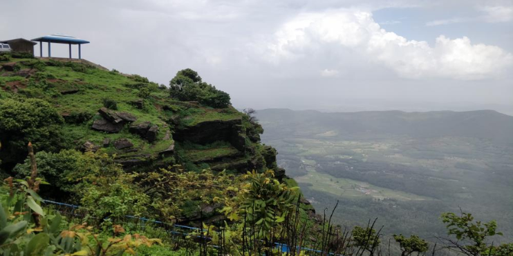 A small hut atop a mountain offers a breathtaking view of the valley, surrounded by lush forests and trekking paths.