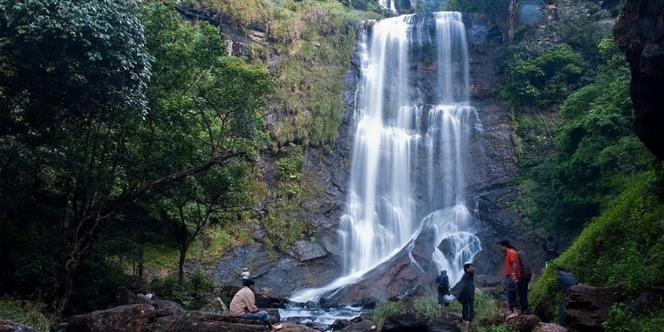 A group of people stands near a stunning waterfall in a lush jungle, enjoying the natural beauty of Hebbe Falls.