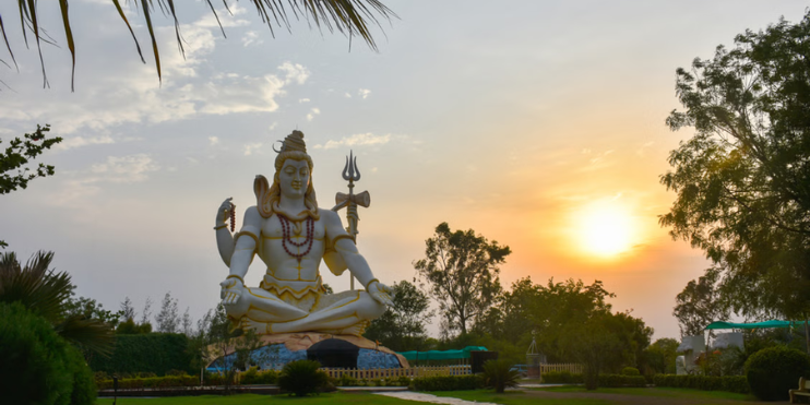 A large statue of Lord Shiva stands majestically in a park, surrounded by lush greenery and serene landscapes.