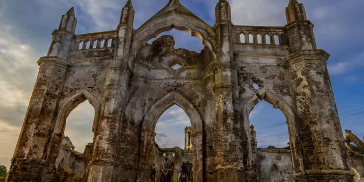 Gothic ruins of Shettihalli Church stand in a field, showcasing pointed arches and ornate stone carvings amidst the landscape.