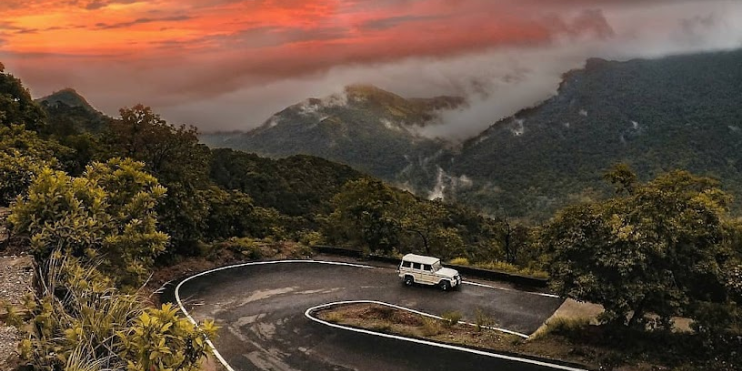 A car navigates a winding mountain road, surrounded by lush greenery and misty peaks in a scenic landscape.