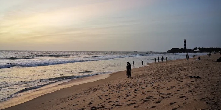People walking along Kapu Beach at sunset, with the old lighthouse silhouetted against the vibrant sky.