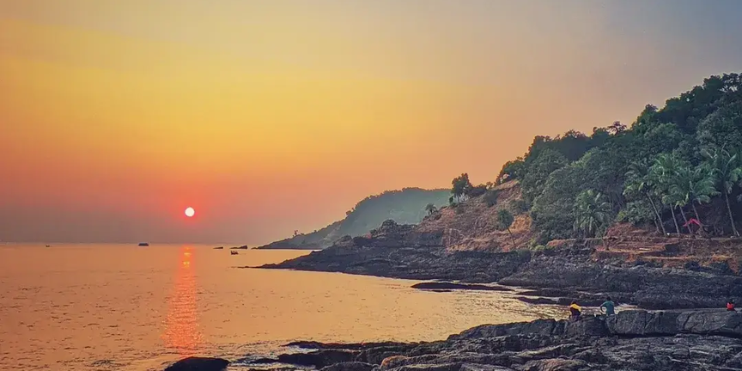 Sunset over Paradise Beach in Goa, showcasing vibrant colors and serene surroundings, framed by rocks and lush greenery.