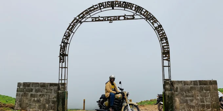 A man sits on a motorcycle under an arch, surrounded by lush greenery, likely near Pushpagiri Wildlife Sanctuary.