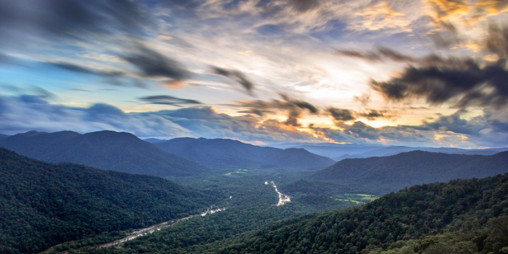 A serene sunset view of mountains and a river, with soft golden light illuminating the landscape and reflecting on the water.